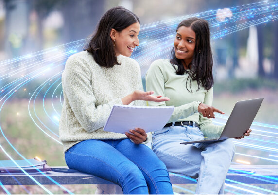 NJIT-ManagedOptical-600x400jpg Young ladies sitting and pointing at laptop while holding a notebook up.