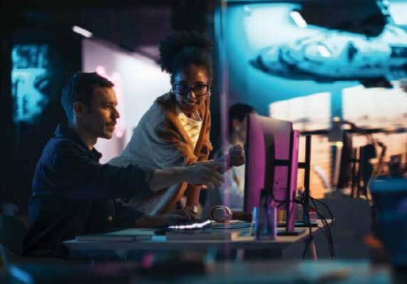 TransformingCampusOperations-600x400 Two colleagues collaborating at a computer in a futuristic, tech-focused workspace with neon lighting and digital displays in the background.